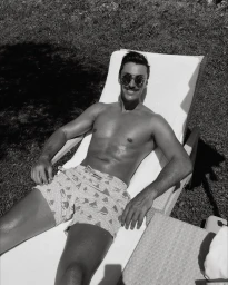 Man wearing sunglasses and patterned swim trunks reclining on an outdoor lounge chair in black and white.