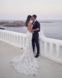 Bride in a white lace wedding dress kissing a groom in a black tuxedo by the ocean with a rocky coastline