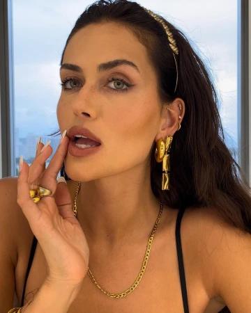 Close-up portrait of a woman with dark hair wearing gold hoop earrings, a chain necklace, and a headband, touching her face in Sydney.