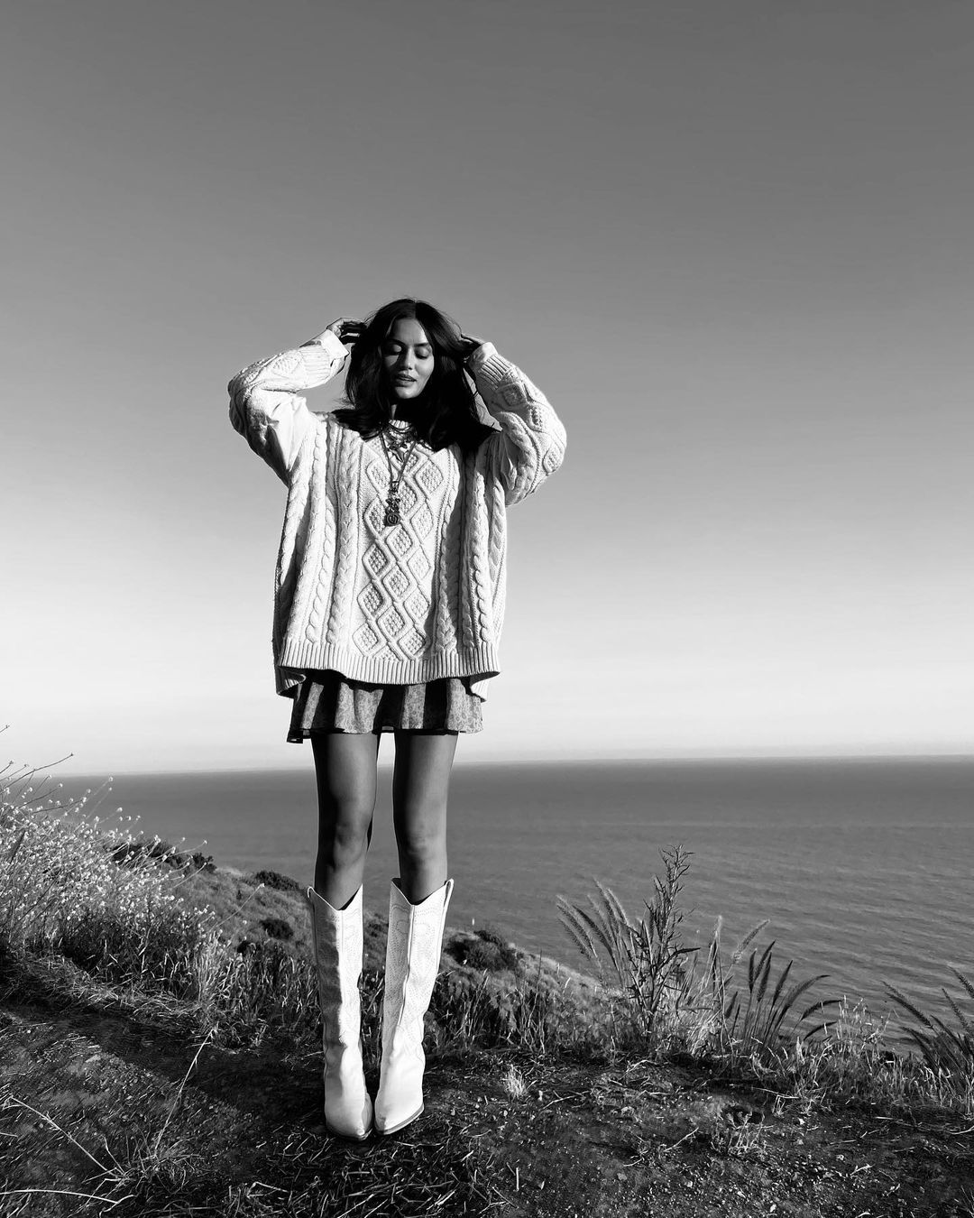 Woman poses on grassy hill overlooking the ocean in oversized sweater, short skirt, and cowboy boots in a black and white outdoor shot.