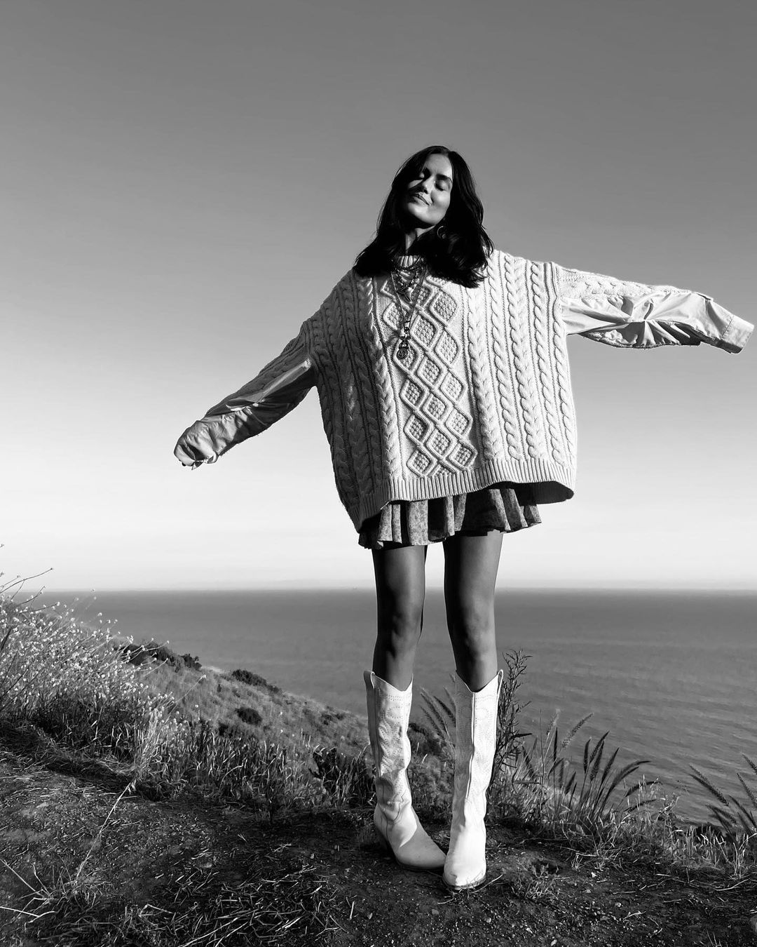 Monochrome full shot of a young woman standing on a grassy hill overlooking the ocean, wearing a sweater and boots with her arms outstretched.