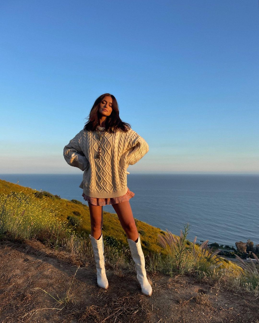 A woman in a sweater, mini dress, and tall boots stands on a grassy hillside overlooking the ocean on a sunny day.