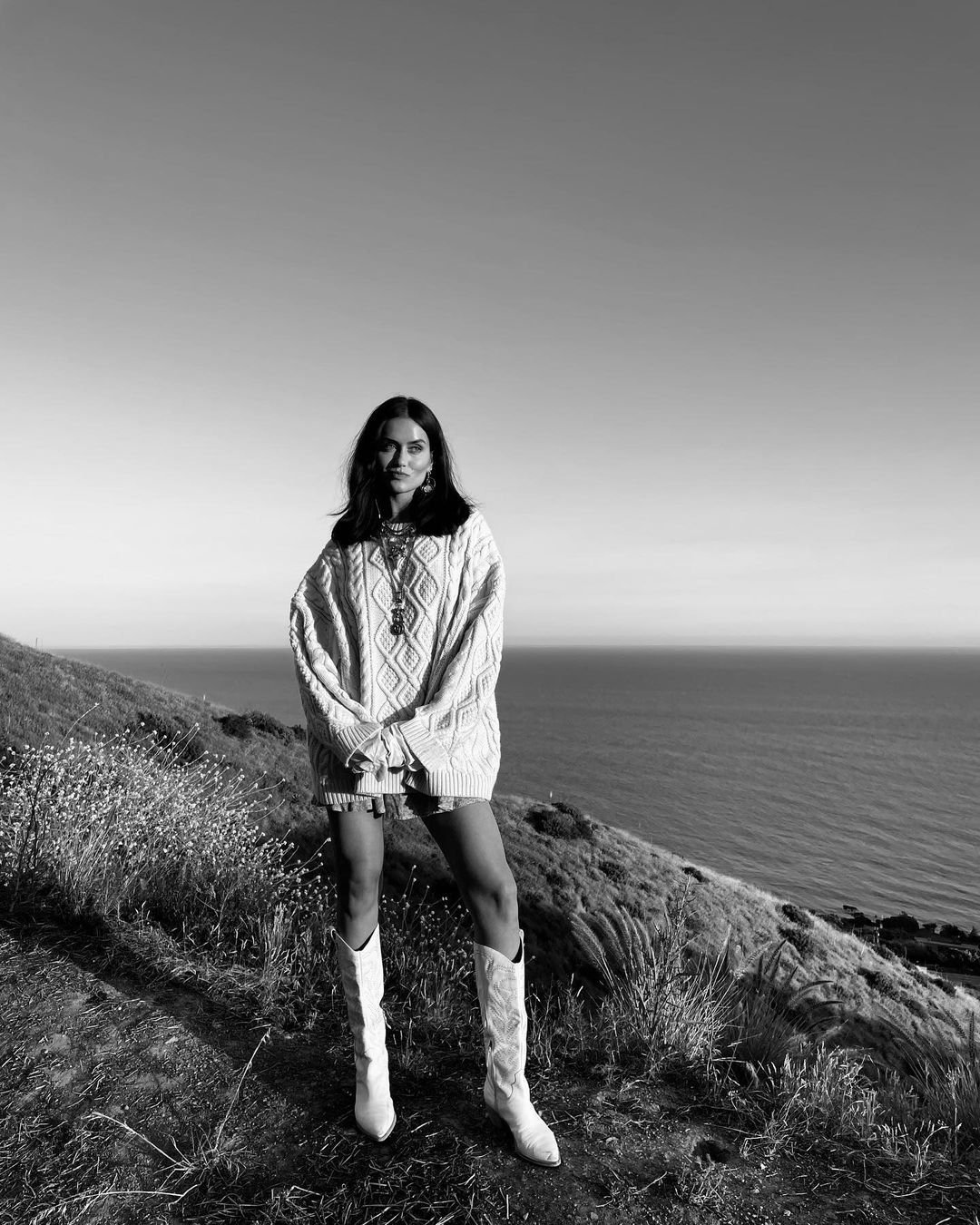 A young woman stands in a sweater dress and boots on a grassy hill overlooking the ocean, photographed in black and white.