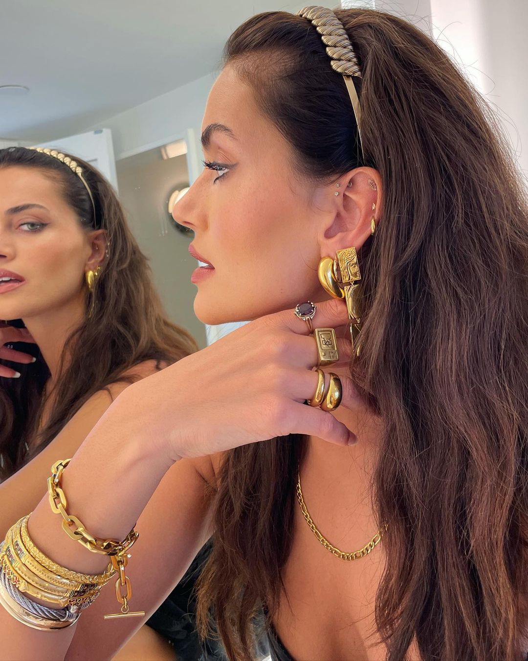 Woman with brunette hair poses indoors with various gold jewelry including rings, bracelets, necklace, and earrings, reflected in a mirror.