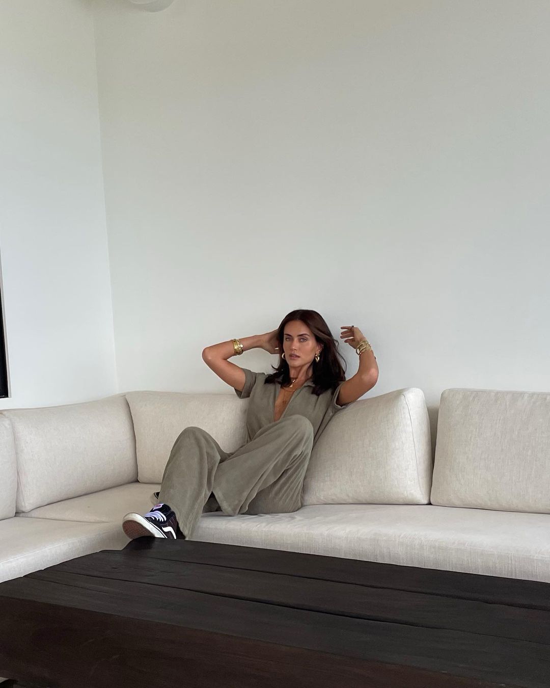 A brunette woman is sitting on a beige couch in a light-filled room, wearing a casual green jumpsuit, sneakers, and gold jewelry.