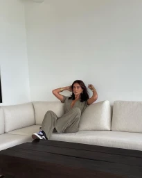 Woman in green jumpsuit sitting on a white sofa with hands behind head by Talia Richman