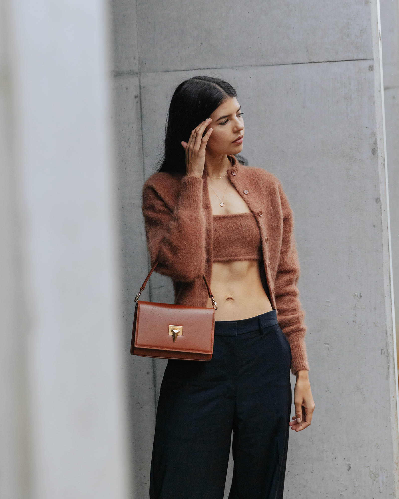 Brunette woman wearing matching knit top and cardigan with wide-leg pants and handbag stands against a textured concrete wall.