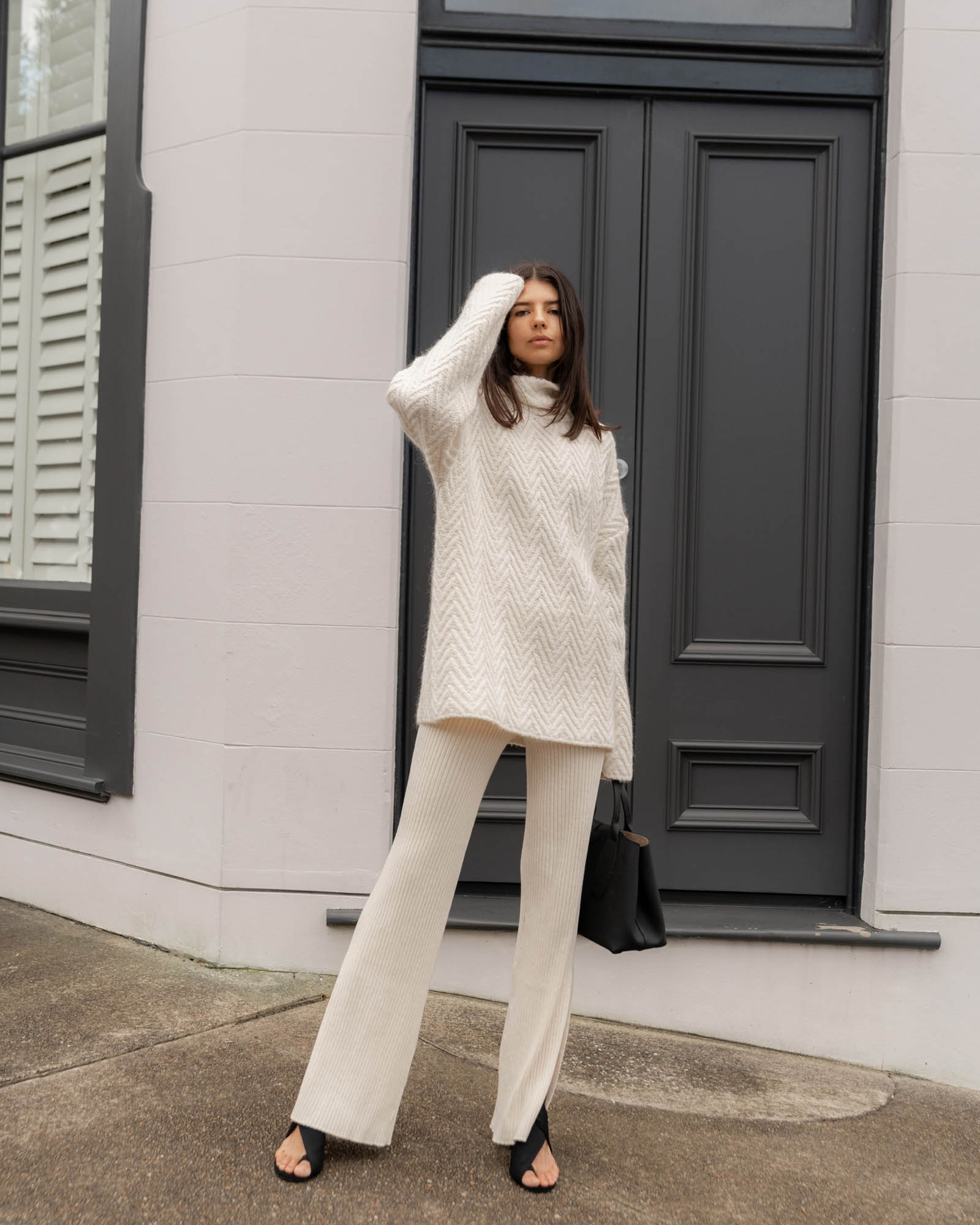 Woman in cream-colored turtleneck sweater and pants standing in front of a dark door with a black tote bag and sandals.