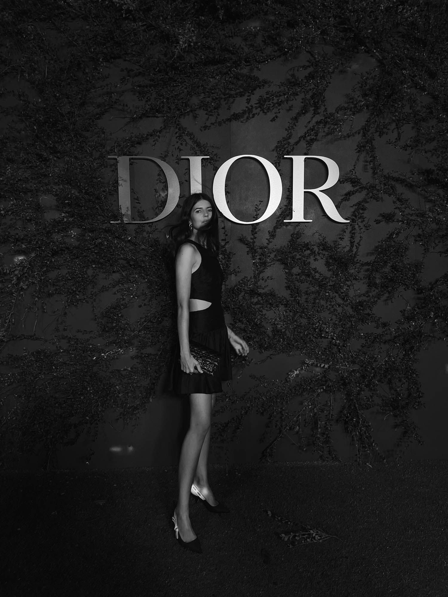 Woman in short black dress with clutch poses in front of ivy wall with 'DIOR' logo in black and white, fashion photography.
