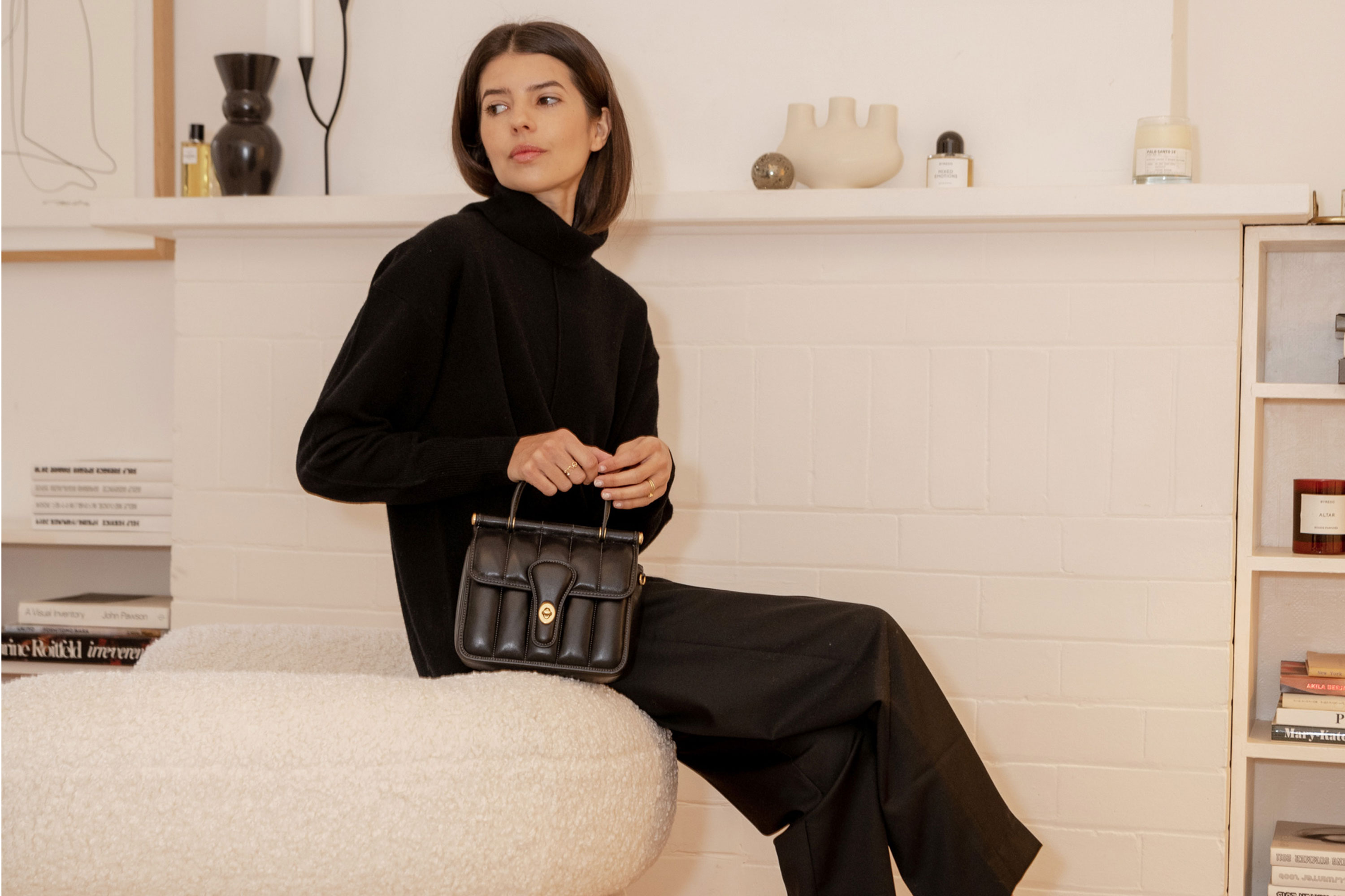 Brunette woman sits in a modern white room wearing a black turtleneck, wide-leg pants, and holding a black leather purse with top handles.
