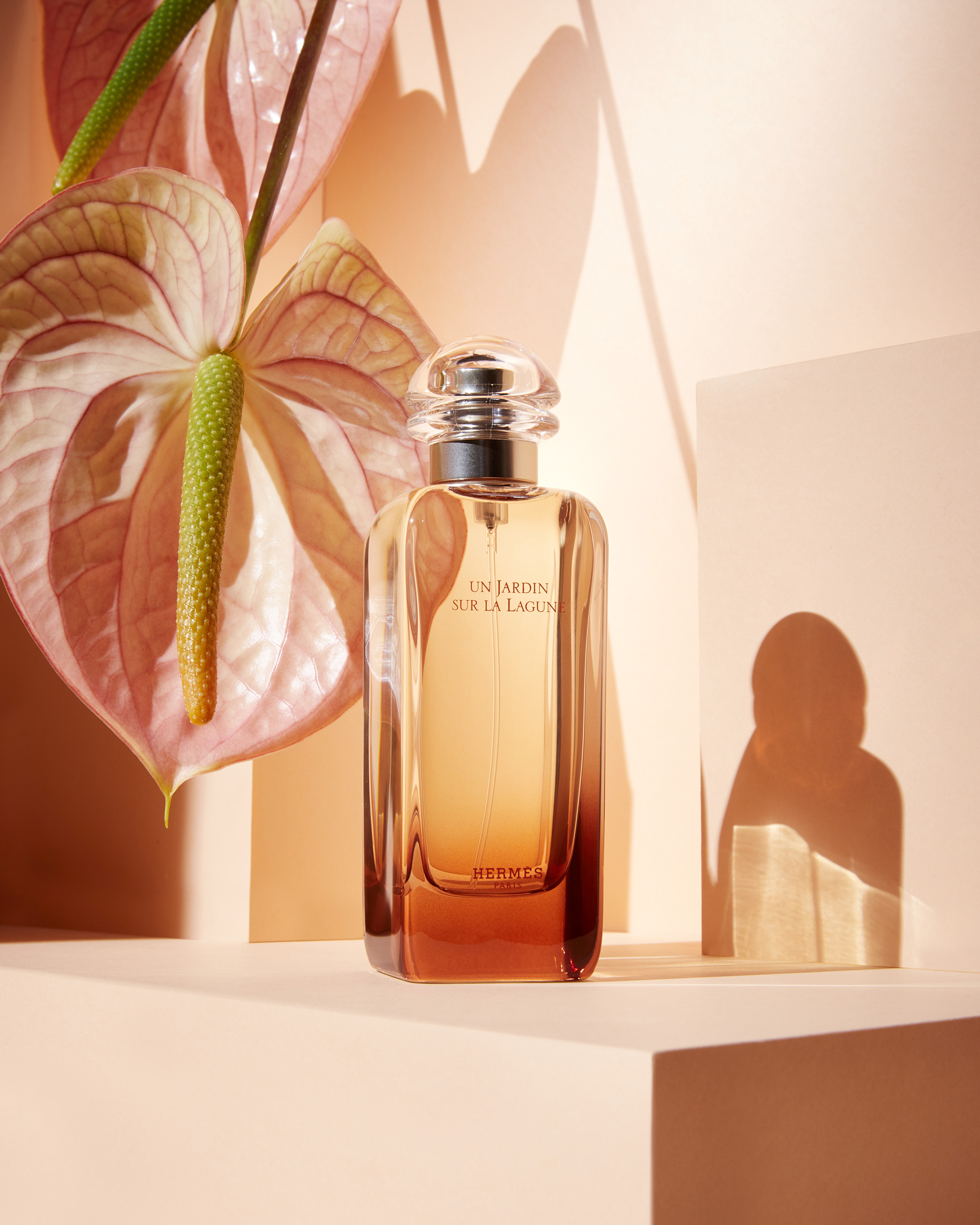 Elegant bottle of Hermes Un Jardin Sur La Lagune perfume alongside a pink Anthurium flower against a peach-toned background.