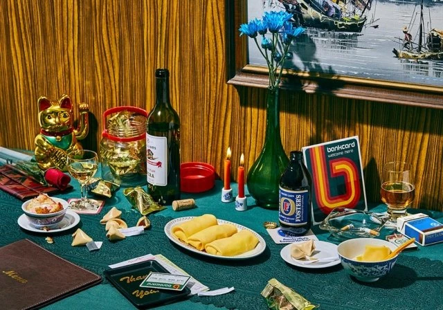Still life with wine bottle, fortune cookies, and vintage props on a table with wood-panel background. Blue flowers in green vase.