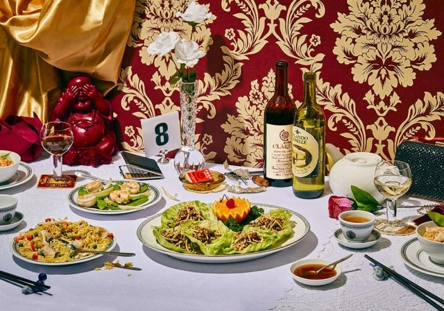 Elaborate still-life food photography featuring assorted Chinese dishes, wine, tea, and decorative elements on a white tablecloth.