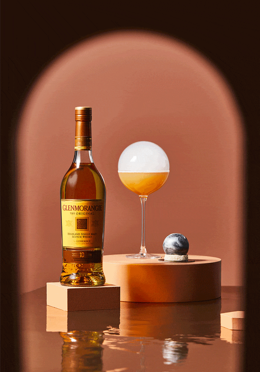Glenmorangie whisky bottle and cocktail glass on peach geometric platforms with water reflection and brown archway background.