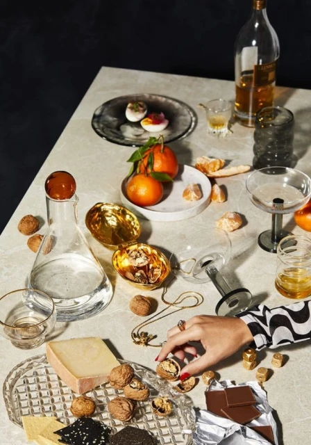 Elegant food and beverage still life featuring cheese, chocolate, walnuts, fruit, and liquor on a textured table surface with a posed hand.