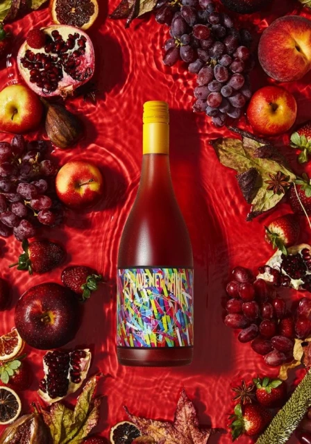 Overhead shot of a red wine bottle with a vibrant label surrounded by colorful fruits and leaves on a rippled red background.
