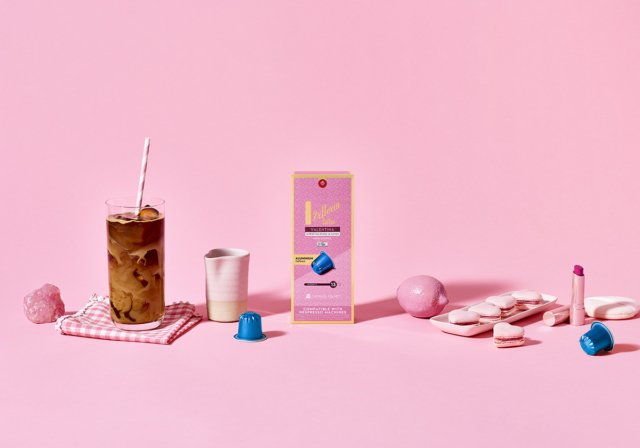Iced coffee in a tall glass, surrounded by coffee capsules, macarons, and a pink aesthetic setting. A creative product flat lay.
