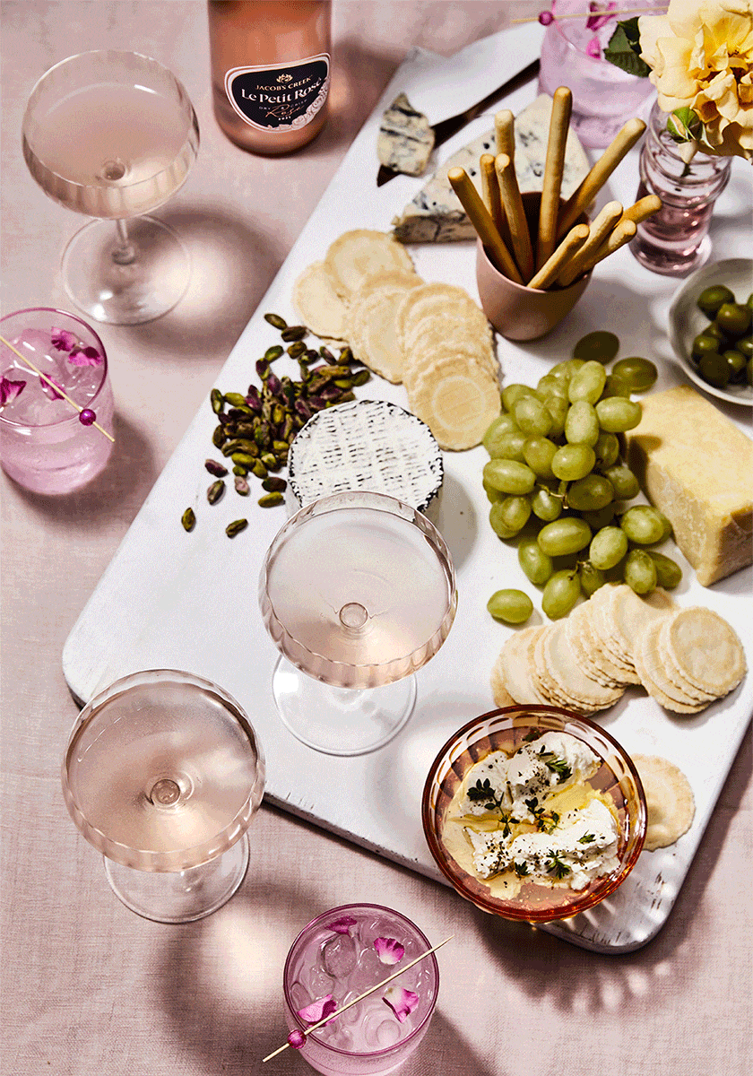 Overhead shot of a white serving board with a variety of cheeses, grapes, olives, pistachios, crackers, cocktails, and flowers.