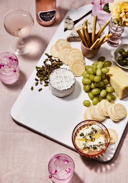 Overhead shot of a white serving board with a variety of cheeses, grapes, olives, pistachios, crackers, cocktails, and flowers.