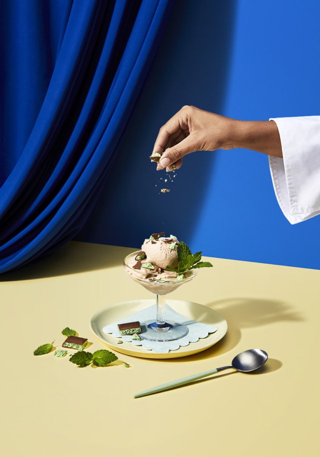 Ice cream sundae with mint leaves being topped with nuts by a hand on a yellow table against a blue curtain backdrop.