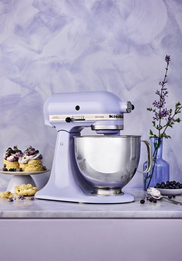Light purple KitchenAid stand mixer with stainless steel bowl, pastries with blueberries, flowers in a vase, and purple background.