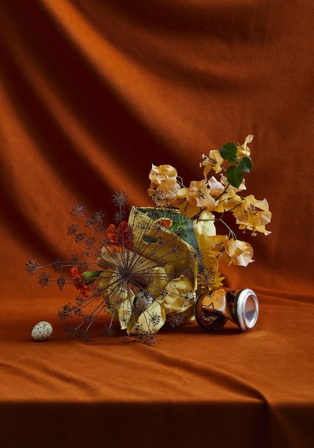 Still life composition featuring a ceramic vase, dried flowers, an egg, and a can on a textured orange fabric background.