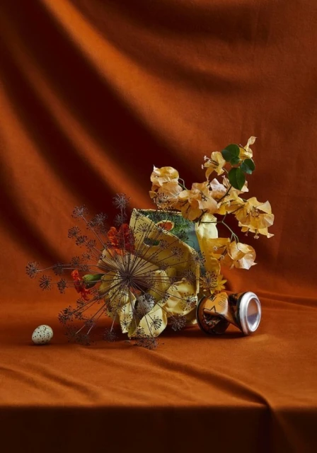Still life composition featuring a ceramic vase, dried flowers, an egg, and a can on a textured orange fabric background.