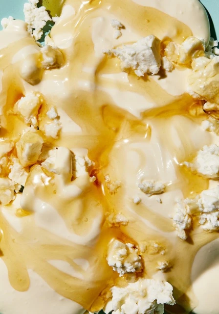 Close-up overhead shot of a dessert with white yogurt, ricotta cheese crumbles, and a drizzle of golden honey on a light blue surface.