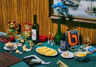 Still life arrangement with a wine bottle, Fosters beer, candles, fortune cookies, and a Bankcard sign on a green tablecloth