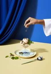 Hand sprinkling chocolate flakes over a dessert in a glass coupe on a yellow surface against a blue backdrop by Tanya Zouev