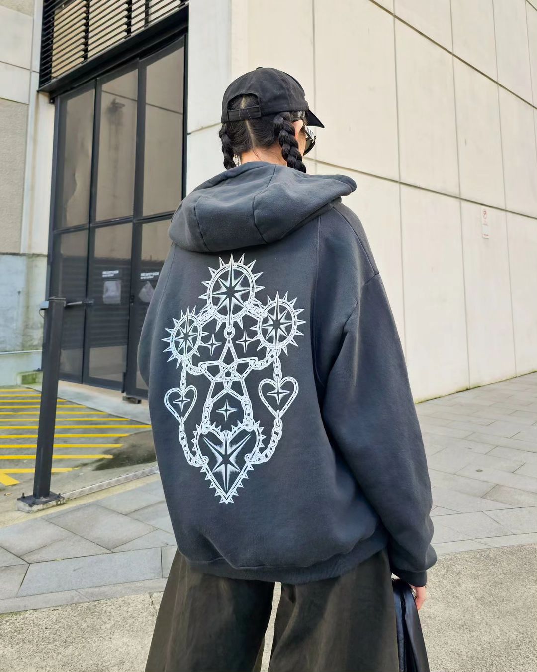 Back view of a person with braided hair wearing a black hoodie with a white graphic design, black hat, and wide-leg olive-green pants outdoors.