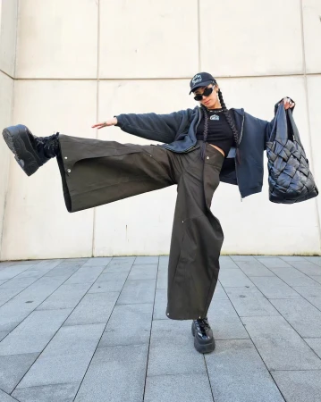Woman in dark zip hoodie, wide olive pants, and platform shoes holding a large black woven bag outside.