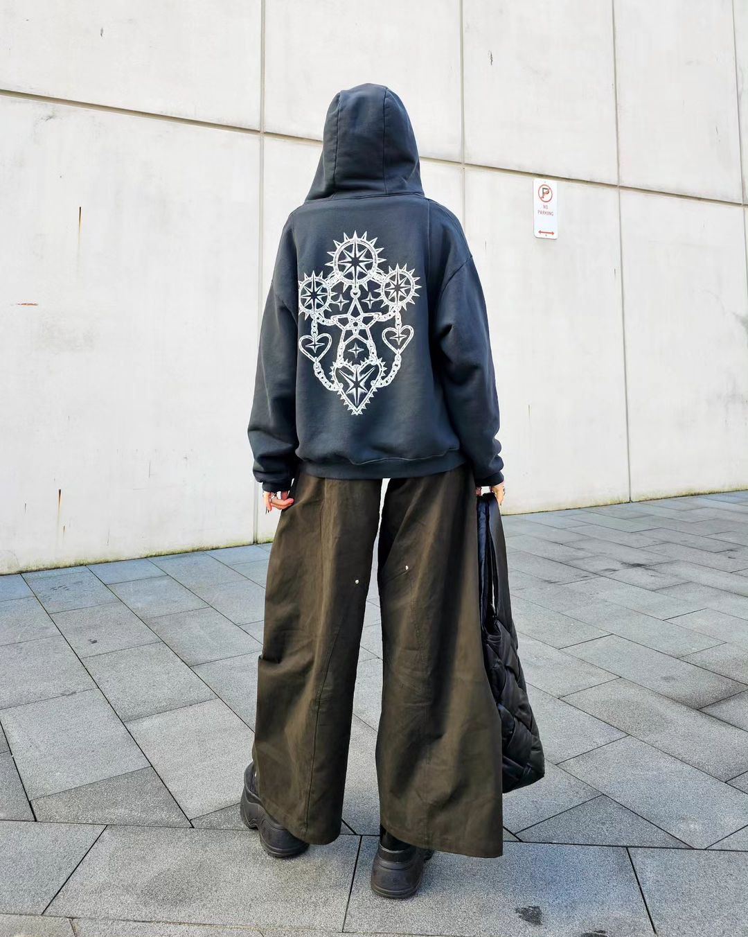 Person in a hooded sweatshirt with a white graphic on the back, wide-legged pants, and platform shoes stands on a sidewalk.