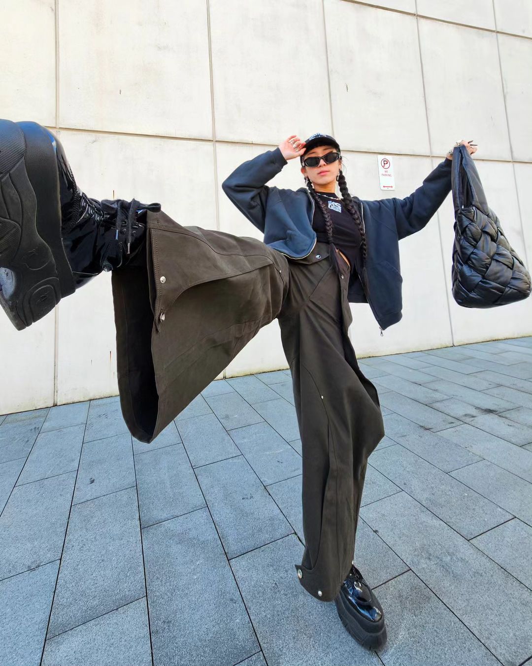 An adult female in a jacket and baggy olive-green pants strikes a pose outside on a sidewalk, holding a black quilted purse and wearing sunglasses.