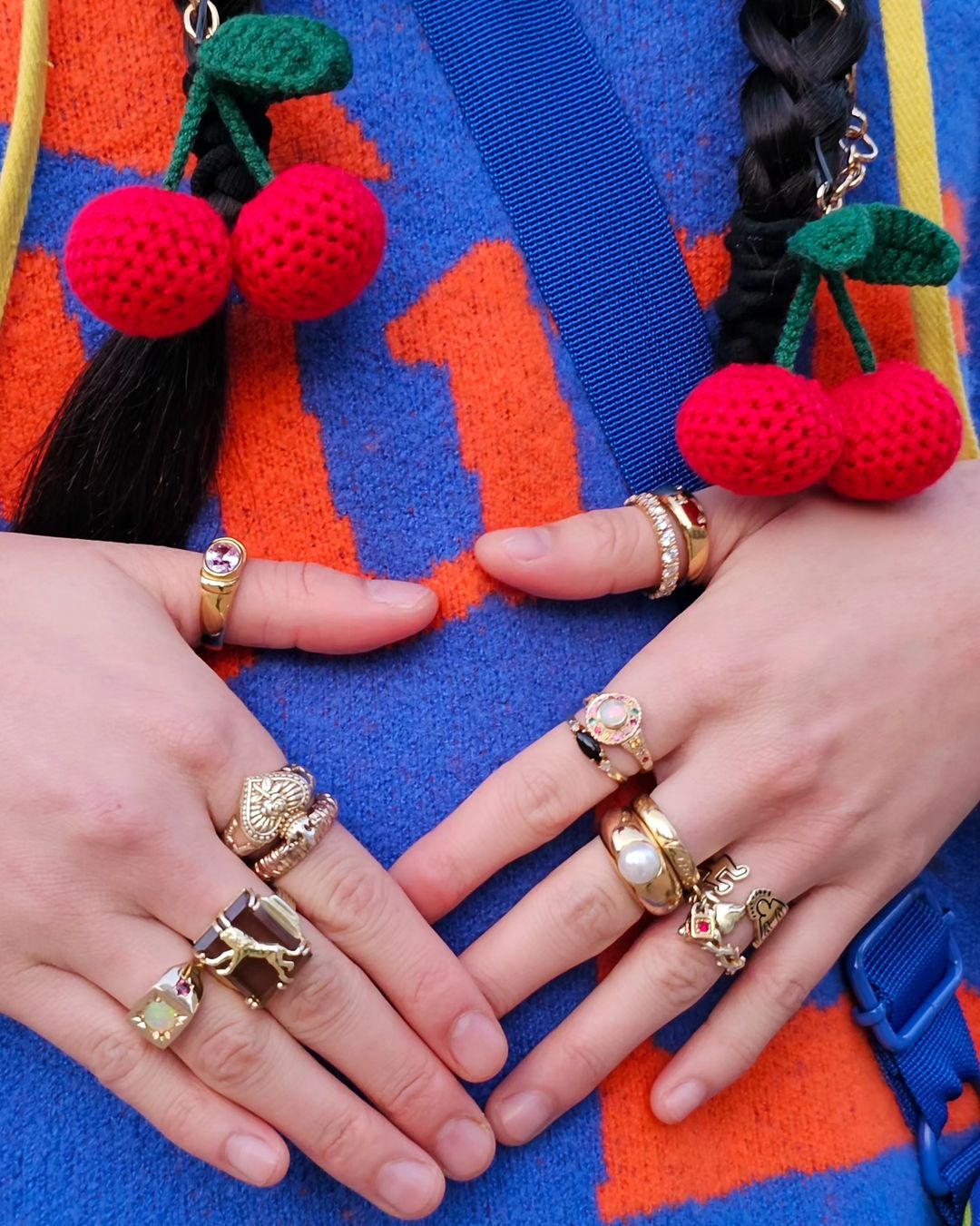 Close-up of woman's crossed hands adorned with numerous rings, showcasing colorful patterned knitwear and hair braids with cherry charms.