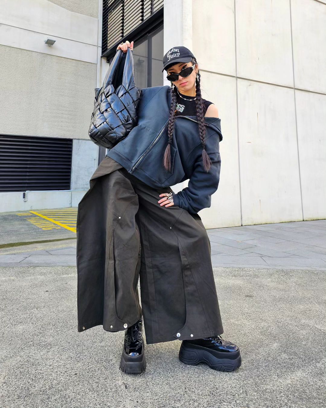 A woman in baggy pants and a jacket poses outdoors, holding a quilted bag. She wears sunglasses and platform boots in an urban setting.
