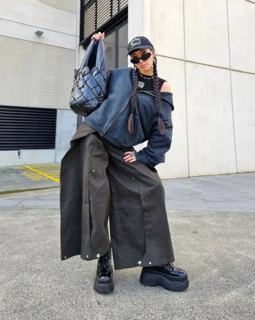 A woman in baggy pants and a jacket poses outdoors, holding a quilted bag. She wears sunglasses and platform boots in an urban setting.