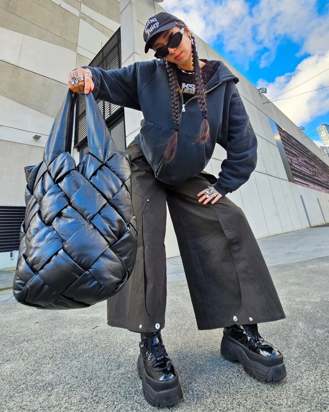 Woman wearing a cap and sunglasses holds a large black woven tote bag against a concrete background, showcasing urban fashion.