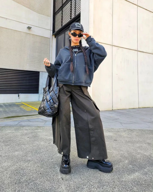 Trendy adult female in a baseball cap, sunglasses, and oversized clothing poses outdoors with a woven tote bag and platform shoes.