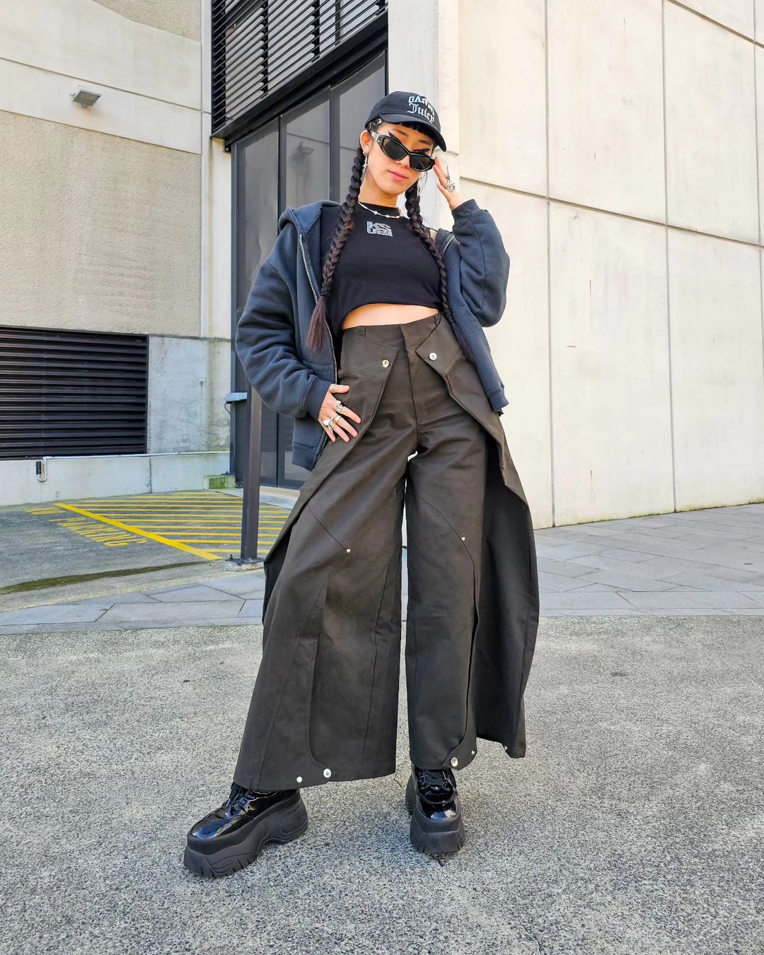Woman in stylish outfit with braids, black crop top, baggy split-leg pants, black platform shoes posing in an urban environment.