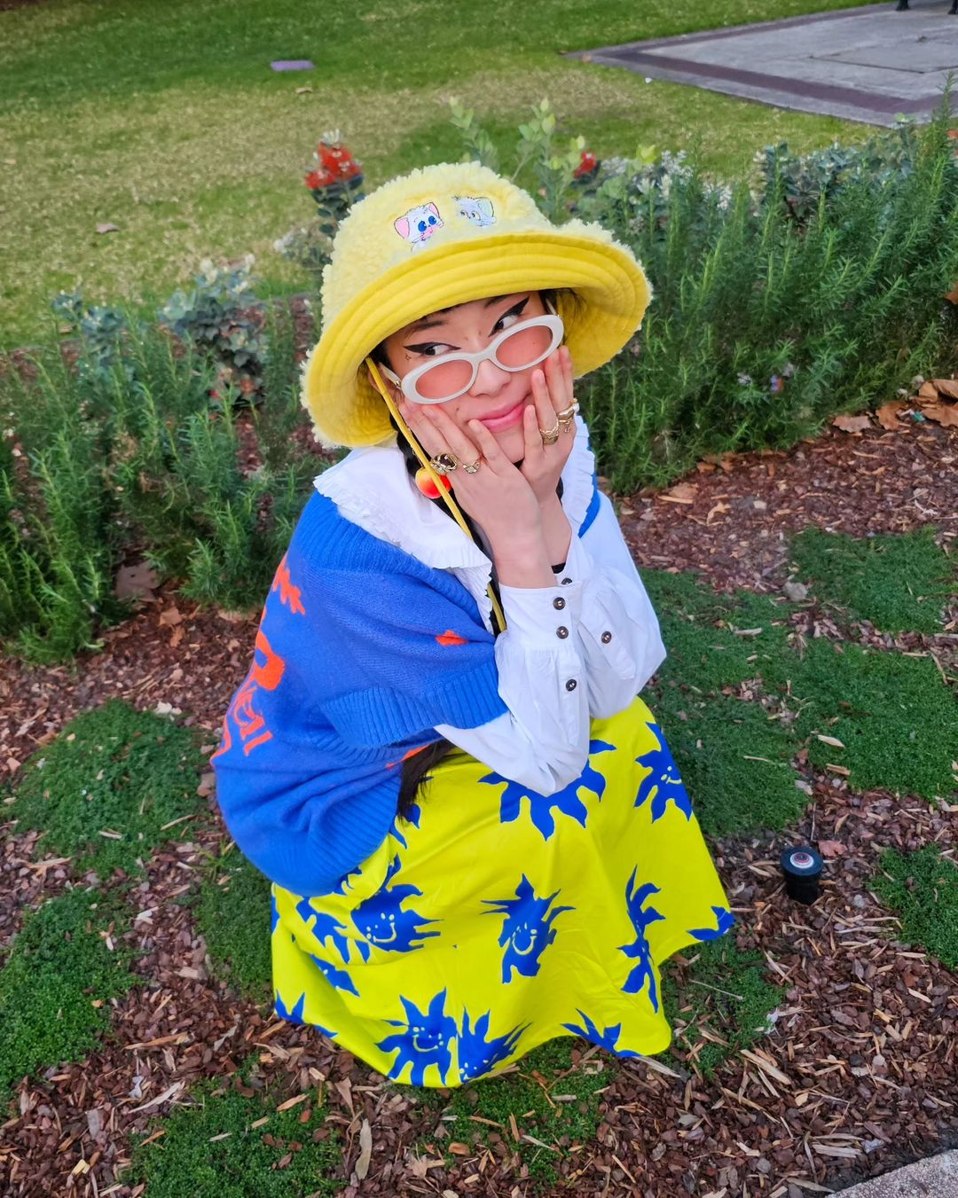 Asian woman in bright yellow hat, skirt with blue sun pattern, and blue sweater sitting on the ground in a park-like setting.