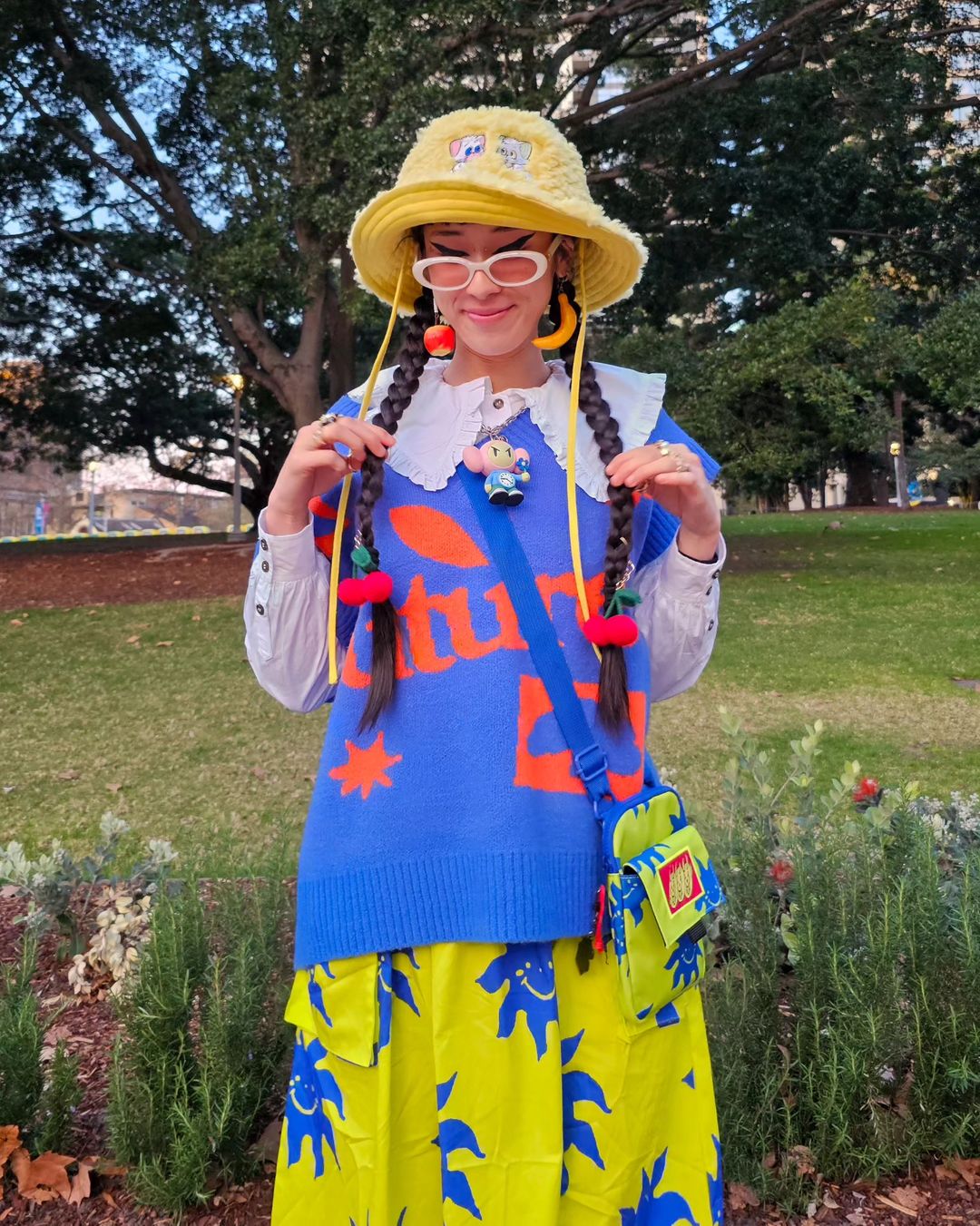 Stylish woman with braids, accessorized with cherries, wears a yellow bucket hat, blue sweater, and yellow skirt in a park setting.