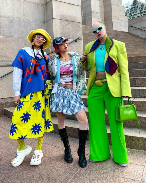 Three people in bright, unique outfits stand on concrete stairs outside a building. Yellows, blues, greens, and plaid fashion statement.