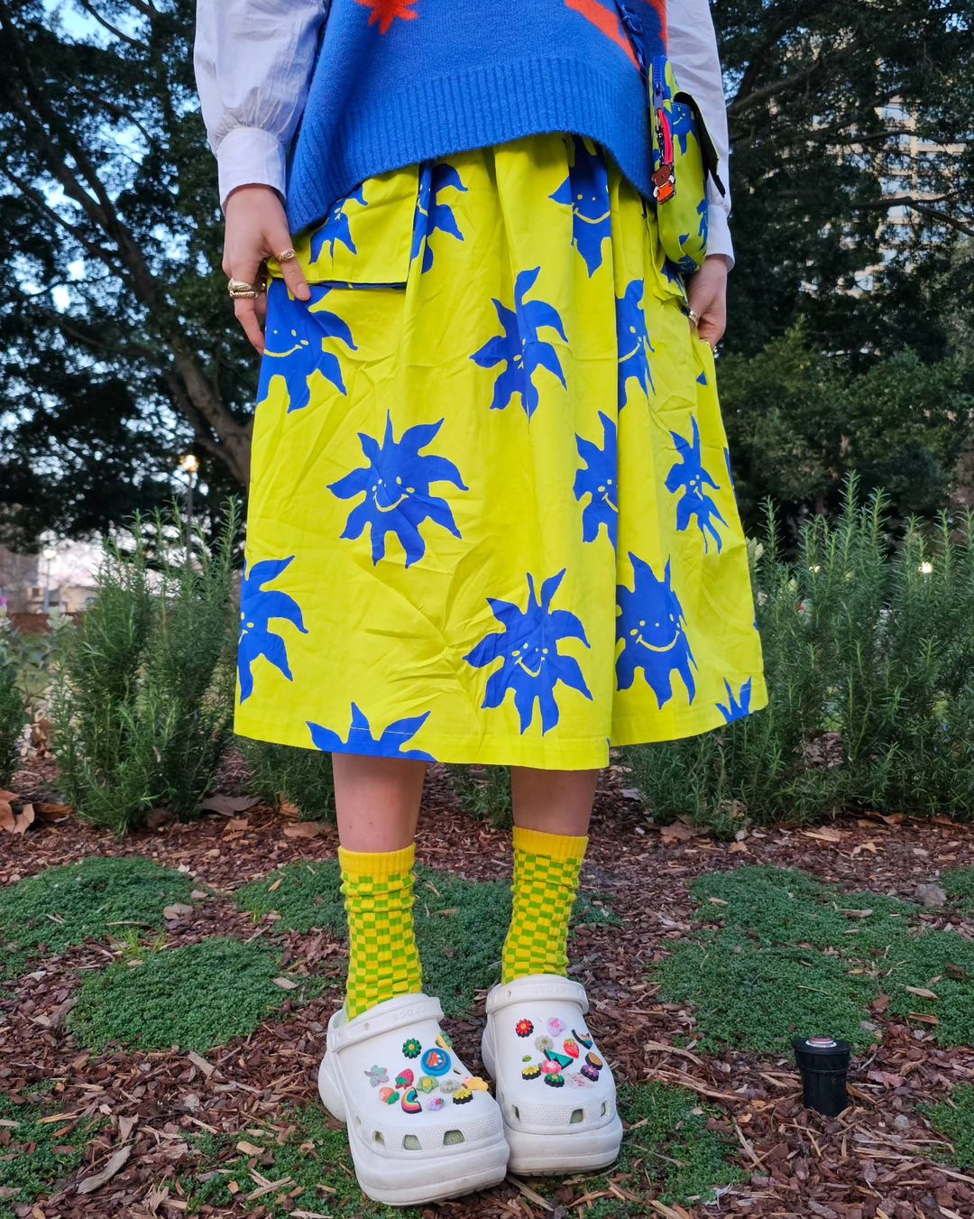 Person standing outdoors wearing a yellow skirt with blue smiling suns, Crocs with charms, and colorful socks.
