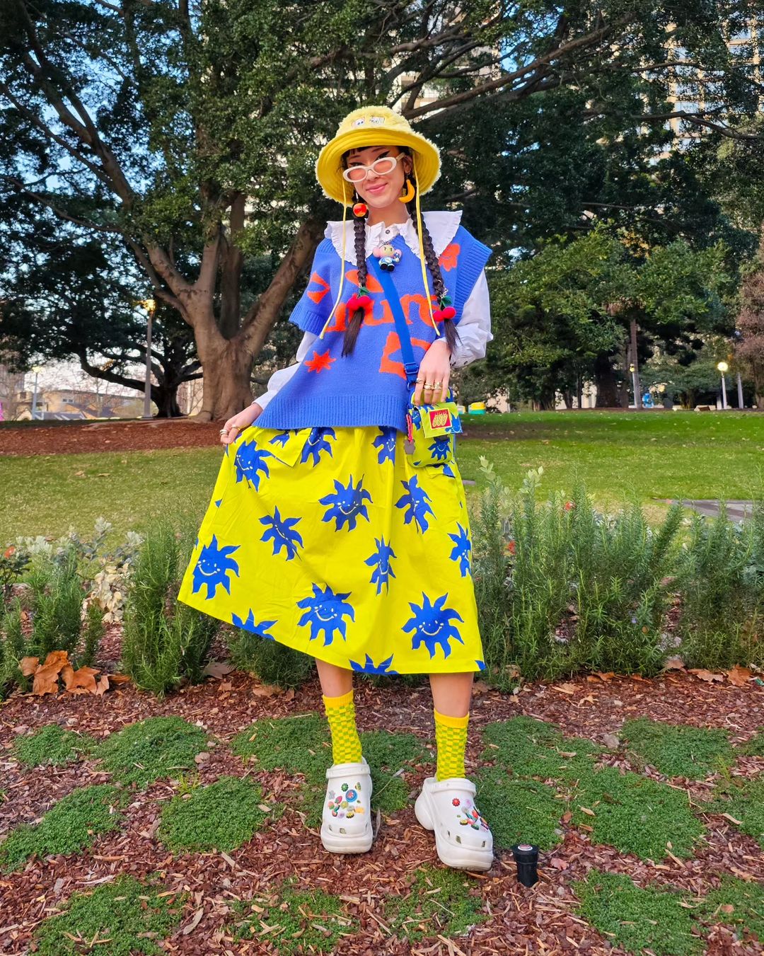 A woman in a yellow hat and colorful outfit stands in a park with greenery and trees in the background, showcasing a fashion-forward look.