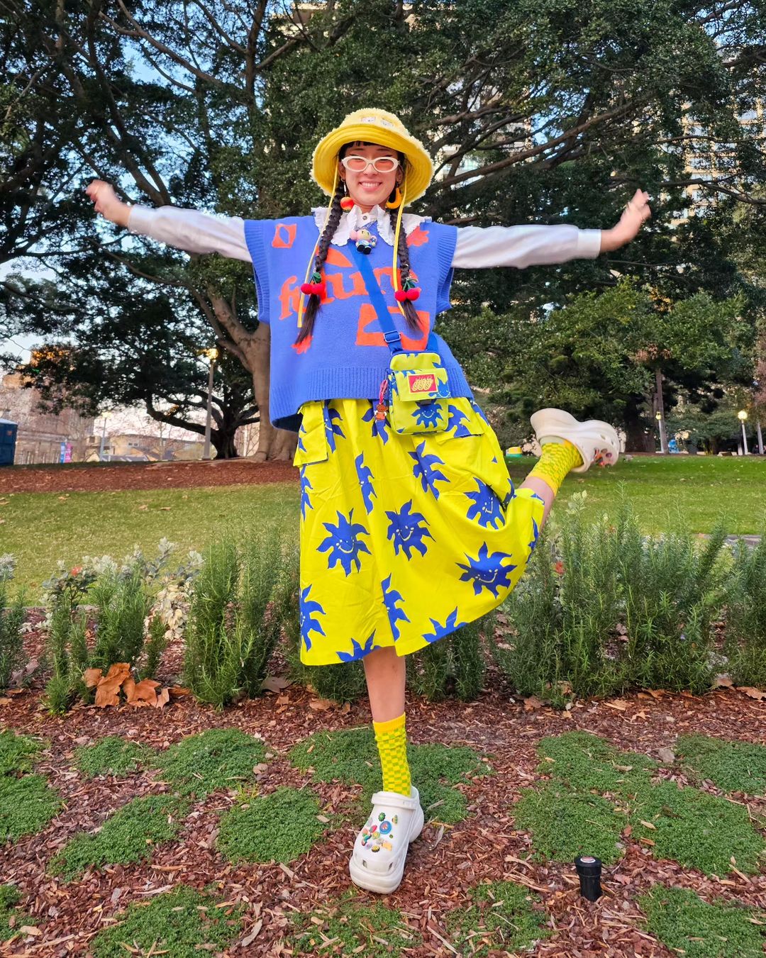 A smiling woman poses in a park, wearing a bucket hat, sweater vest, printed skirt, novelty socks, and platform shoes for a fun, colorful fashion look.