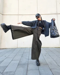 Woman in dark zip hoodie, wide olive pants, and platform shoes holding a large black woven bag outside.