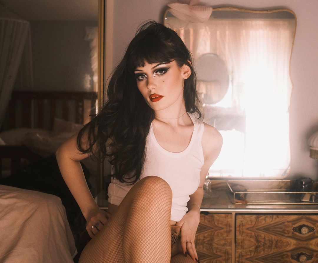 A young adult female poses indoors, wearing a white tank top and fishnet stockings, with dark hair and bold makeup, seated by a wooden dresser.