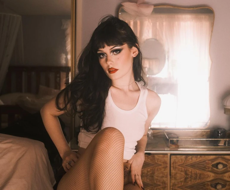 A young adult female poses indoors, wearing a white tank top and fishnet stockings, with dark hair and bold makeup, seated by a wooden dresser.