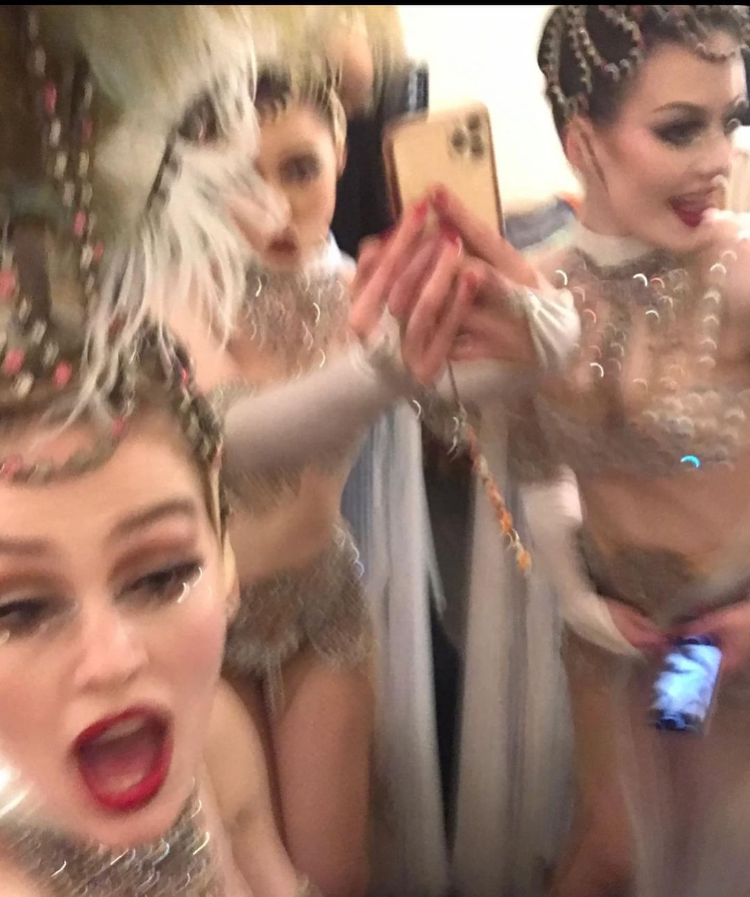 Three adult females in costumes take a selfie; two are visible, the other holds a phone. All have elaborate hair and makeup; indoor setting.